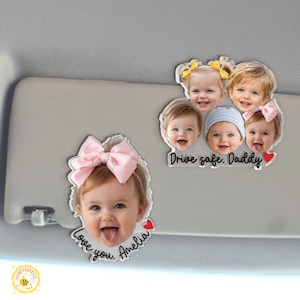 May include: Clear acrylic car visor ornaments featuring smiling baby faces. One ornament has a pink bow and the text "Love you, Amelia" with a red heart. Another has multiple baby faces and the text "Drive safe, Daddy" with a red heart.