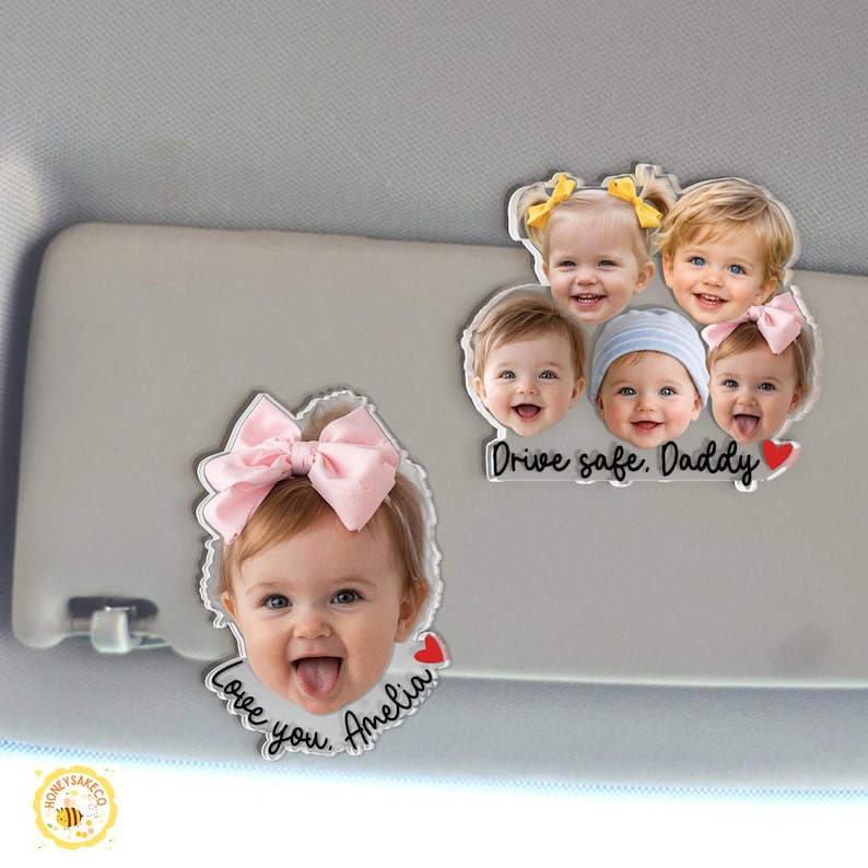 May include: Clear acrylic car visor ornaments featuring smiling baby faces. One ornament has a pink bow and the text "Love you, Amelia" with a red heart. Another has multiple baby faces and the text "Drive safe, Daddy" with a red heart.