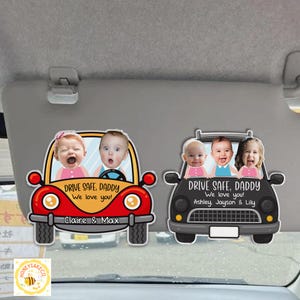May include: Two car-shaped sun visor ornaments. One is red with a yellow top, the other is black. Each features a photo of children and the text "DRIVE SAFE, DADDY We love you!" with names below. A cute and personalized car accessory.