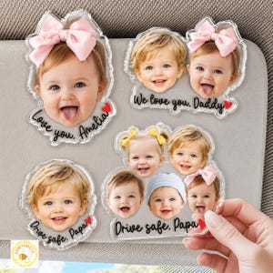 May include: Clear acrylic car visor magnets featuring smiling baby faces. Some magnets have pink bows, and text that reads "Love you, Amelia", "We love you, Daddy", and "Drive safe, Papa".
