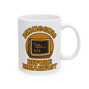 May include: White ceramic mug with the words "DEBUGGING BEFORE BREAKFAST" in orange lettering. The mug features a retro-style design with a computer screen graphic in a brown circle.