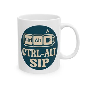 Puede incluir: Taza de cerámica blanca con un gráfico azul y beige. El gráfico presenta el texto "Ctrl Alt Sip" con teclas de teclado y un icono de taza de café. La taza tiene un asa curva y es adecuada para bebidas calientes.