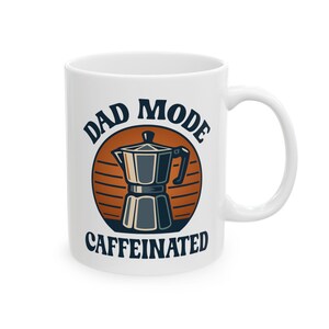 Op de afbeelding: Witte keramische koffiemok met de woorden "DAD MODE CAFFEINATED" in een cirkelvormig ontwerp. Het ontwerp bevat een illustratie van een koffiezetapparaat in blauw en zwart, tegen een oranje en bruin gestreepte achtergrond.
