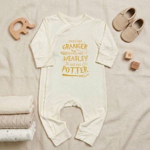May include: Cream-colored baby onesie with long sleeves and the text "Study like Granger, Protect like Weasley, Live like Potter" in gold. The image includes a wooden elephant toy, baby shoes, and blocks.