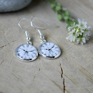 May include: A pair of silver dangle earrings with a white clock face featuring a black silhouette of the Eiffel Tower. The clock face has Roman numerals.