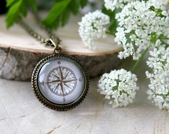 May include: A bronze compass pendant necklace with a white background and black compass markings. The necklace is on a wooden surface with white flowers.