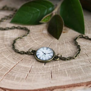 Puede incluir: Un brazalete de cadena de bronce con un colgante redondo en forma de esfera de reloj. La esfera es blanca con números romanos negros y agujas negras.