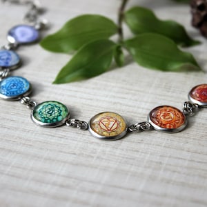 Op de afbeelding: Een zilveren kettingarmband met zeven ronde bedels met de zeven chakrasymbolen in hun overeenkomstige kleuren. De bedels zijn gerangschikt in een regenboogpatroon, beginnend met paars en eindigend met rood.