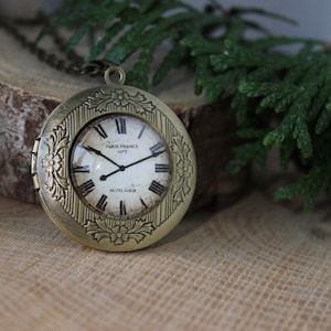 May include: A gold-toned locket necklace with a clock face that reads "Paris France 1973" and "Heures Vapeur". The locket is decorated with floral details and has a hinged opening.