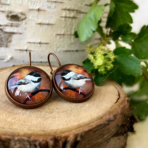 Pode incluir: Um par de brincos de cobre com um charme redondo de vidro que apresenta uma imagem pintada de um chapim-real pousado em um galho. O fundo do charme é de cor laranja e marrom quente.