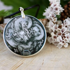 May include: A silver pendant necklace with a round, black and white image of a dragon. The dragon is depicted in a swirling, cloud-like design.