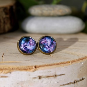 Op de afbeelding: Een paar bronzen oorbel studs met een galaxy-thema ontwerp. De oorbellen hebben een ronde, heldere glazen cabochon met een draaiende patroon van paars, blauw en wit.