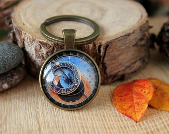 May include: A bronze-colored key chain with a round glass charm featuring a detailed replica of the Prague Astronomical Clock. The clock face is blue, orange, and gold with Roman numerals and black markings.