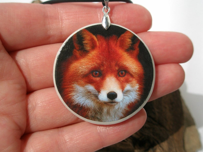 Op de afbeelding: Een ronde hangerketting met een close-up afbeelding van een rode vos met witte vacht op zijn borst. De vos heeft een zwarte achtergrond en zijn ogen kijken rechtstreeks naar de kijker.