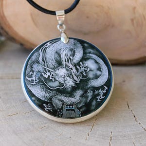 May include: A silver pendant necklace with a black cord. The pendant is a round, white and black cameo featuring a detailed carving of a dragon.
