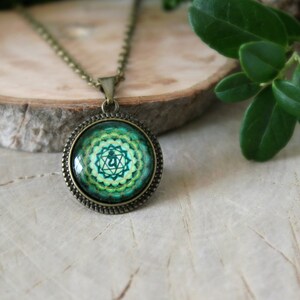 May include: A green and gold necklace with a round pendant featuring a green and gold mandala with a symbol in the center. The necklace is on a wooden surface.