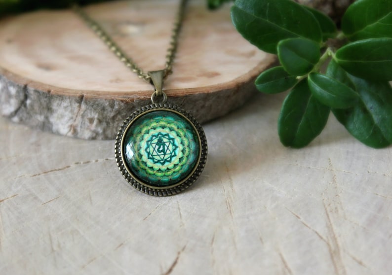 May include: A green and gold necklace with a round pendant featuring a green and gold mandala with a symbol in the center. The necklace is on a wooden surface.