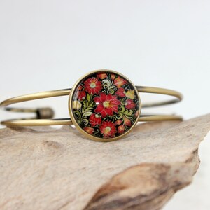 Op de afbeelding: Een gouden armband met een ronde cabochon met een rood, geel en groen bloemenontwerp.