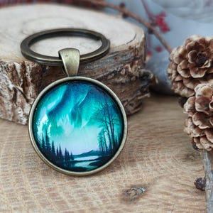 May include: A round glass keychain with a bronze metal frame. The glass features a painting of a forest scene with a lake and a green and blue aurora borealis in the sky.