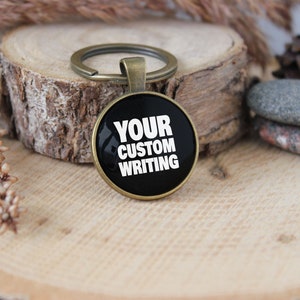 Peut inclure: Un porte-clés noir et bronze avec un charme rond en verre qui dit "YOUR CUSTOM WRITING" en texte blanc.