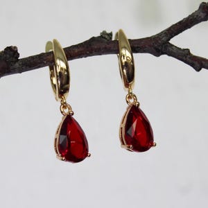 May include: A pair of gold-coloured hoop earrings, each with a teardrop-shaped, faceted, red gemstone. The earrings are suspended from a brown twig against a soft, neutral background. The gemstones have a deep red colour.