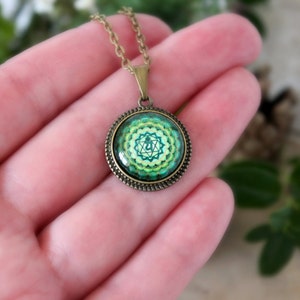 May include: A green and gold necklace with a round pendant featuring a green and yellow mandala with a symbol in the center. The pendant is surrounded by a gold-colored frame.
