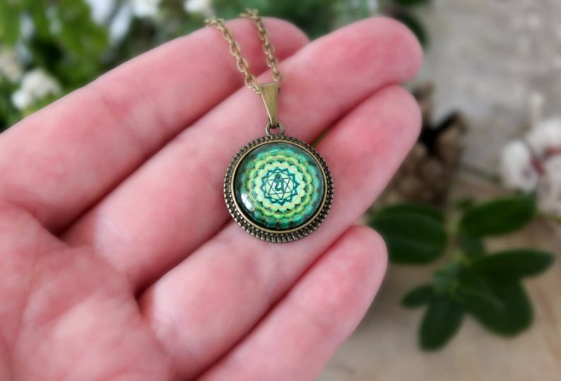 May include: A green and gold necklace with a round pendant featuring a green and yellow mandala with a symbol in the center. The pendant is surrounded by a gold-colored frame.