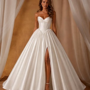 May include: A strapless white satin wedding gown with a sweetheart neckline and a high slit. The full skirt flows to the floor. The model is wearing white strappy heels. The background is a neutral color.