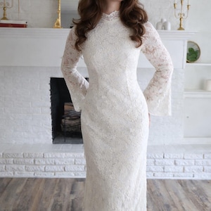 May include: A long, ivory lace dress with a high neckline and flared sleeves. The dress is floor-length and has a fitted bodice. The model is standing in front of a white brick fireplace.