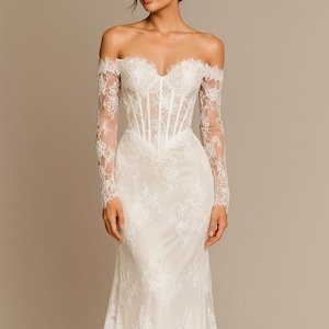 May include: Off-the-shoulder ivory wedding gown with long sleeves. The dress features a fitted bodice with vertical boning and a flowing skirt. Delicate lace detailing covers the sleeves and bodice, adding texture and elegance to the bridal attire.