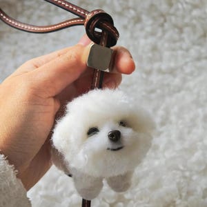 Pode incluir: Um pingente de cachorro Bichon Frise branco e fofo com uma alça de couro marrom e fecho de metal. O cachorro tem uma expressão sorridente e olhos e nariz pretos pequenos. O pingente é preso a uma alça de couro marrom com um nó.