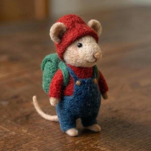 May include: A handcrafted felt mouse figurine wearing a red knit cap, red shirt, blue overalls, and a green backpack. The mouse has tan ears and a long, pale tail. The figurine is standing on a wooden surface.