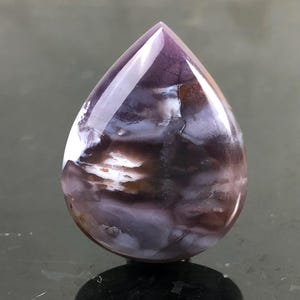 May include: A polished, teardrop-shaped gemstone with a mix of colors. The stone features a deep purple at the top, transitioning to white, brown, and lavender hues. The surface is smooth and reflective, showcasing the natural patterns within.
