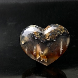 May include: A heart-shaped stone with a polished, translucent surface. The stone features a mix of brown, black, and gold inclusions, creating a unique pattern. The heart is displayed against a dark background.