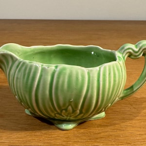 May include: A light green ceramic creamer with a scalloped rim and a decorative handle. The creamer has a ribbed texture and sits on three small feet. The overall design is vintage and elegant.