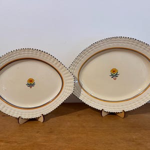 May include: Two oval serving platters. The cream-coloured surfaces feature a yellow floral design. The rims have a woven texture and are edged with black dots. The platters are supported by wooden stands. Measurements are in centimetres.