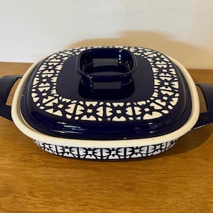 May include: A square enamelled metal casserole dish with a matching lid. The exterior and lid feature a dark navy blue base with a repeating white geometric pattern. The dish has two integrated side handles and a square knob on the lid.