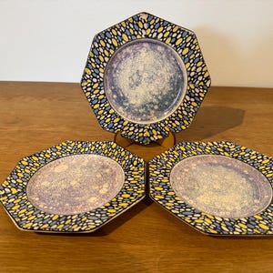 May include: Three decorative plates with an octagonal design. The plates have a speckled border in yellow, blue, and black, framing a central design in purple and white. They are displayed on a wooden surface.