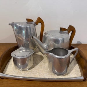 May include: A vintage tea set comprising a teapot, creamer, sugar bowl, and a serving tray. The set is made of brushed aluminium with wooden handles. The tray has a light-coloured woven insert and a dark wood border.