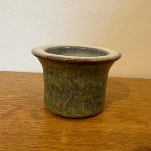 May include: A small, handcrafted ceramic pot with a speckled green and brown glaze. The rim is a lighter beige colour, and the pot has a slightly flared shape. The pot is sitting on a wooden surface.
