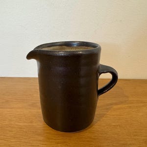 May include: A dark brown ceramic jug with a glossy finish, featuring a small pouring spout and a side handle. The interior of the jug has a light beige, speckled texture, while the exterior surface shows subtle horizontal ridges.