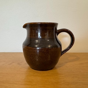 May include: A brown ceramic jug with a rounded body, a spout, and a curved handle. The jug has a glossy finish and sits on a wooden surface. The dark brown colour gives it a rustic, handcrafted appearance. The jug is likely used for serving beverages.