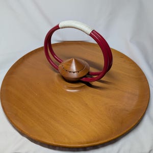 May include: A circular wooden serving tray with a burgundy handle and a wooden knob. The handle is wrapped with white material. The tray is made of light brown wood and has a smooth surface. The tray is designed for serving food or displaying items.