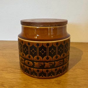 May include: A cylindrical ceramic coffee canister featuring a glossy brown glaze and a dark brown geometric pattern. The word COFFEE is embossed in a band around the centre. The container is topped with a circular, flat, dark brown wooden lid.