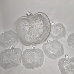 May include: A collection of clear glass apple-shaped serving bowls. The large bowl and individual bowls feature a textured surface and a clear stem detail. The bowls are displayed on a white surface.