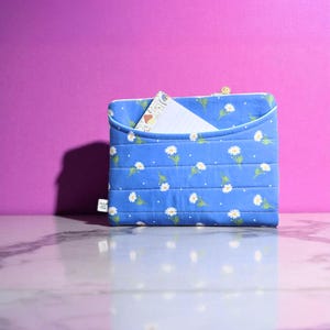 May include: Blue quilted laptop sleeve with a daisy pattern. The sleeve has a pocket and a zipper closure. A notepad is visible in the pocket. The background is purple.