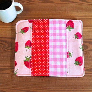 May include: A rectangular, quilted placemat with a patchwork design. The placemat features sections of pink fabric with strawberry patterns, red polka dots, and pink gingham. A white coffee mug sits in the upper left corner, on a wooden surface.
