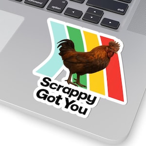 May include: A sticker featuring a brown rooster with a red comb and wattle, set against a backdrop of colorful stripes. The text "Scrappy Got You" is below the rooster. The sticker is placed on a laptop.