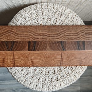 May include: A rectangular wooden cutting board with a natural wood grain pattern. The board is made of three horizontal sections of varying shades of brown. The board is resting on a cream-colored, woven surface.