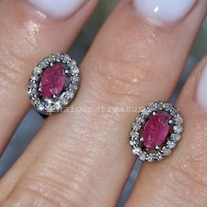 May include: A pair of oval stud earrings. Each earring has a vibrant red gemstone surrounded by a halo of small, sparkling white stones. The earrings are set in a silver-coloured metal. The background is a hand with white nail polish.